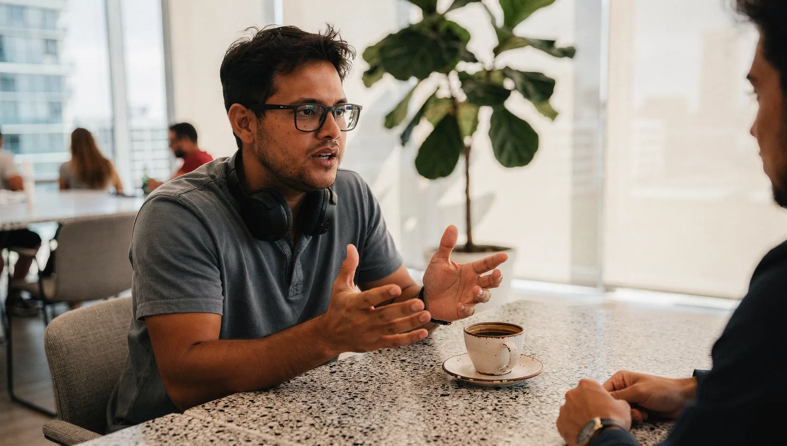Man with glasses and headphones around his neck gestures while talking at a terrazzo table in a sunlit office.
