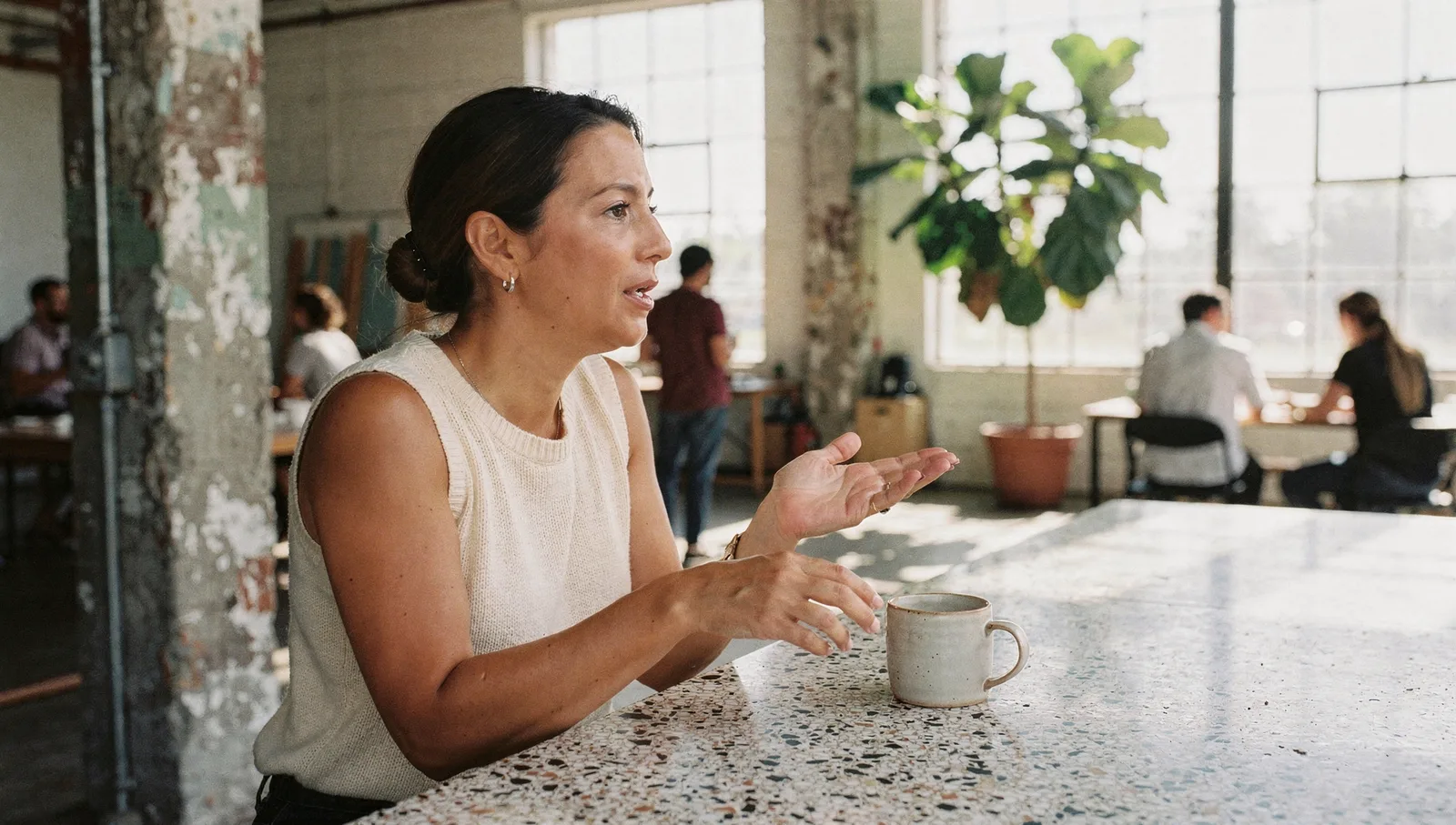 A woman in a cream top gestures while speaking, holding a ceramic mug in a sunlit warehouse studio.