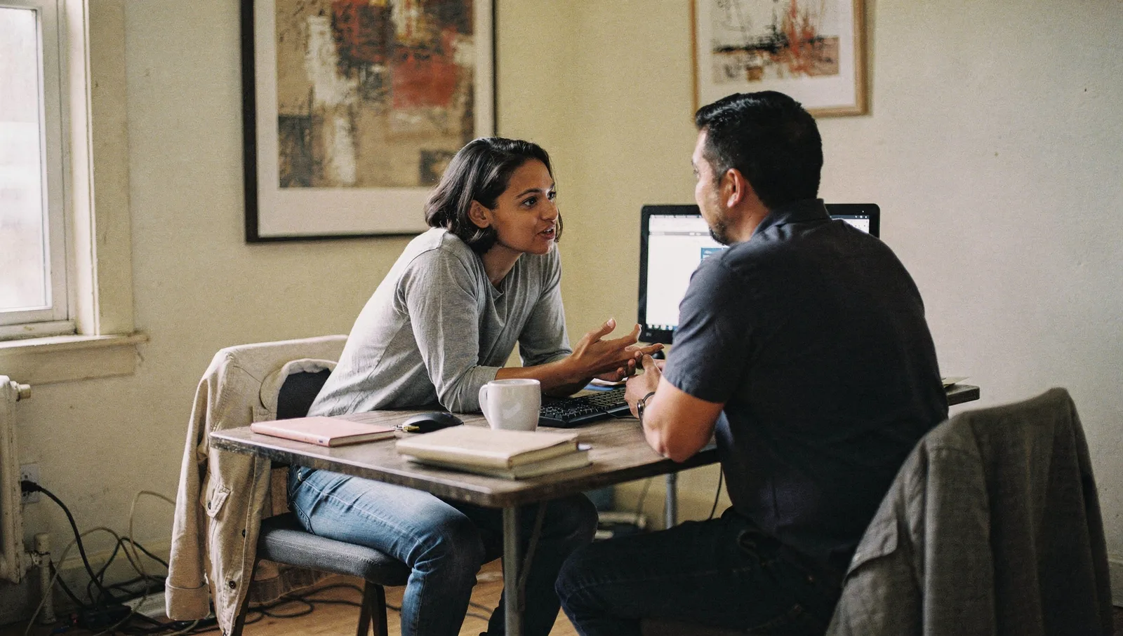 Two colleagues sit at a desk in a sunlit office, conversing naturally during a collaborative work session.
