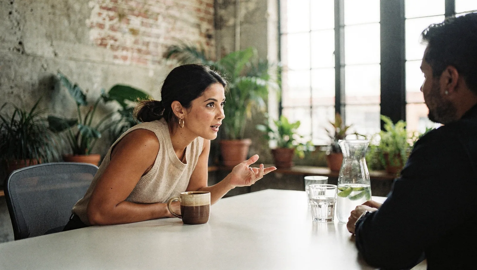 Two colleagues conversing across a conference table in a sunlit meeting room with exposed concrete and indoor plants.