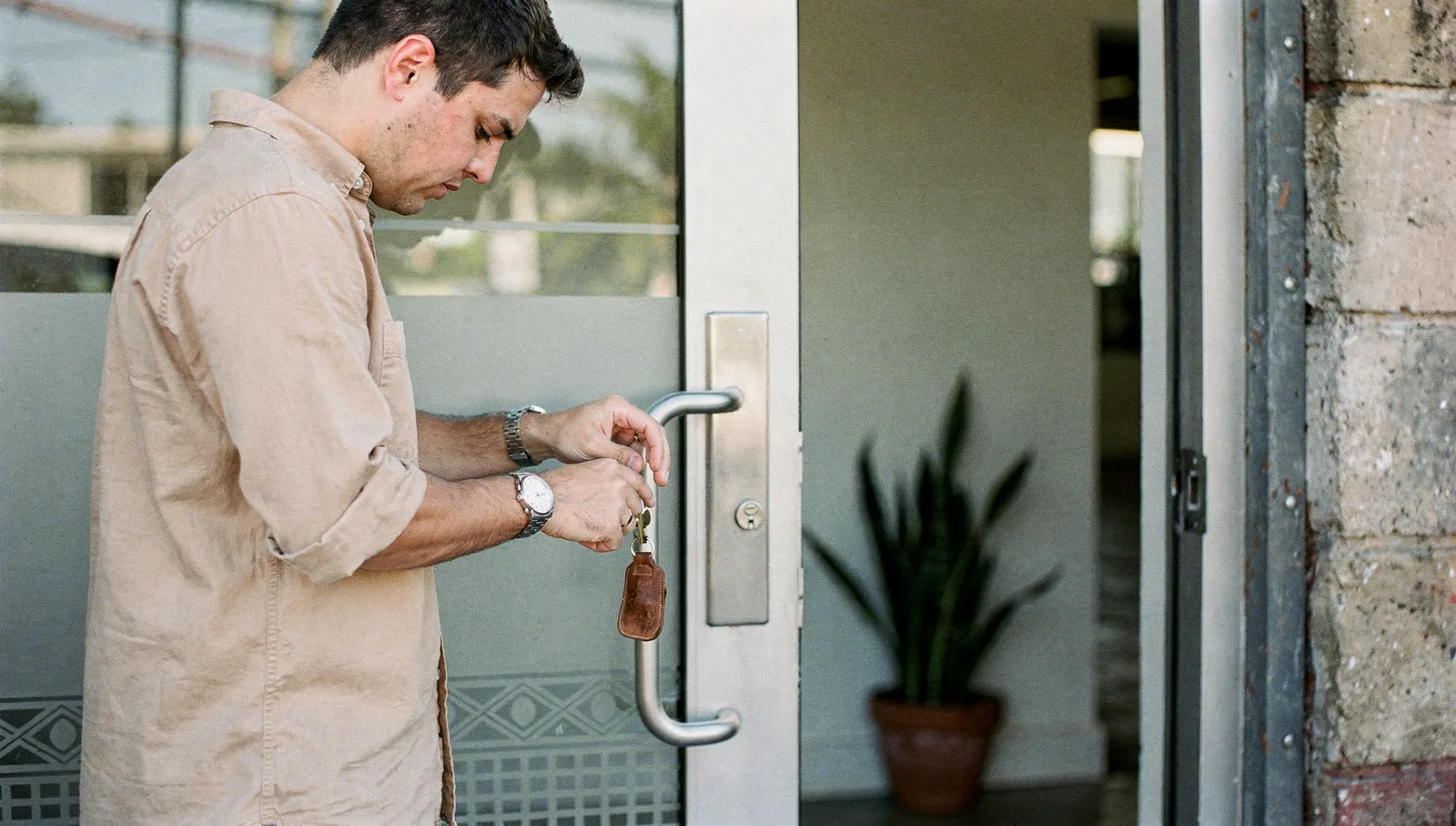 A person unlocking the glass door of an office building in morning sunlight.