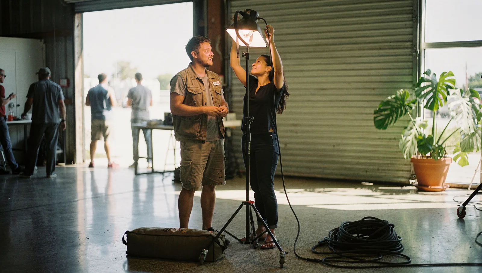Two people adjust studio lighting and gear inside a sunlit warehouse for a marketing photoshoot.