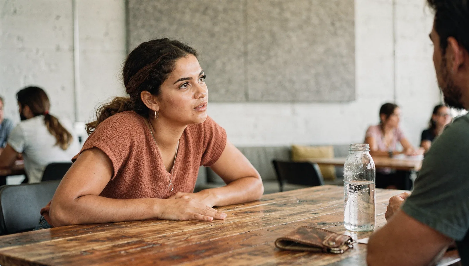 A woman in a terracotta top leans over a wood table, listening intently during a meeting in a sunlit loft office.