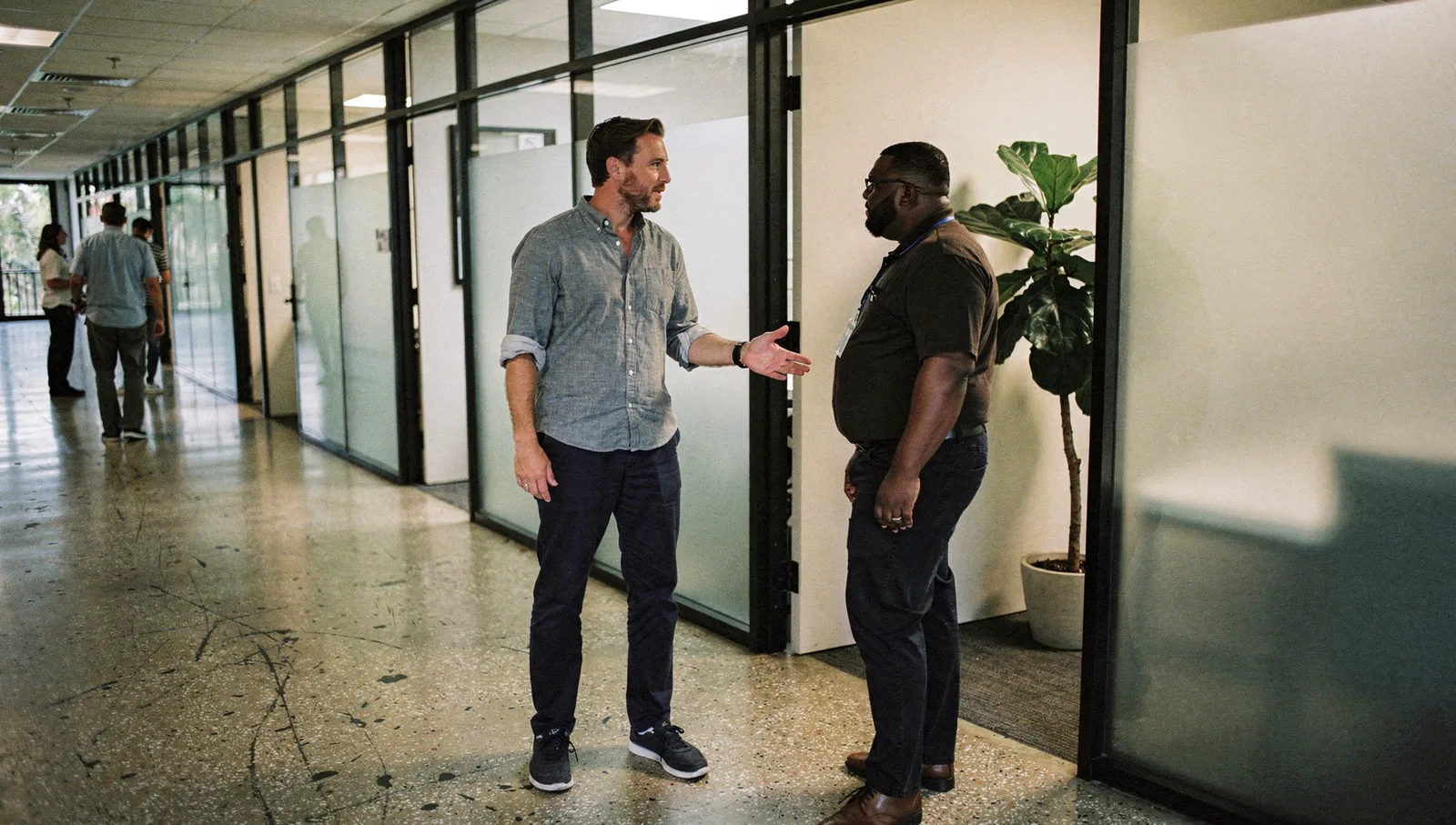 Two professionals converse in a glass-partitioned office hallway with a polished concrete floor and a potted plant.