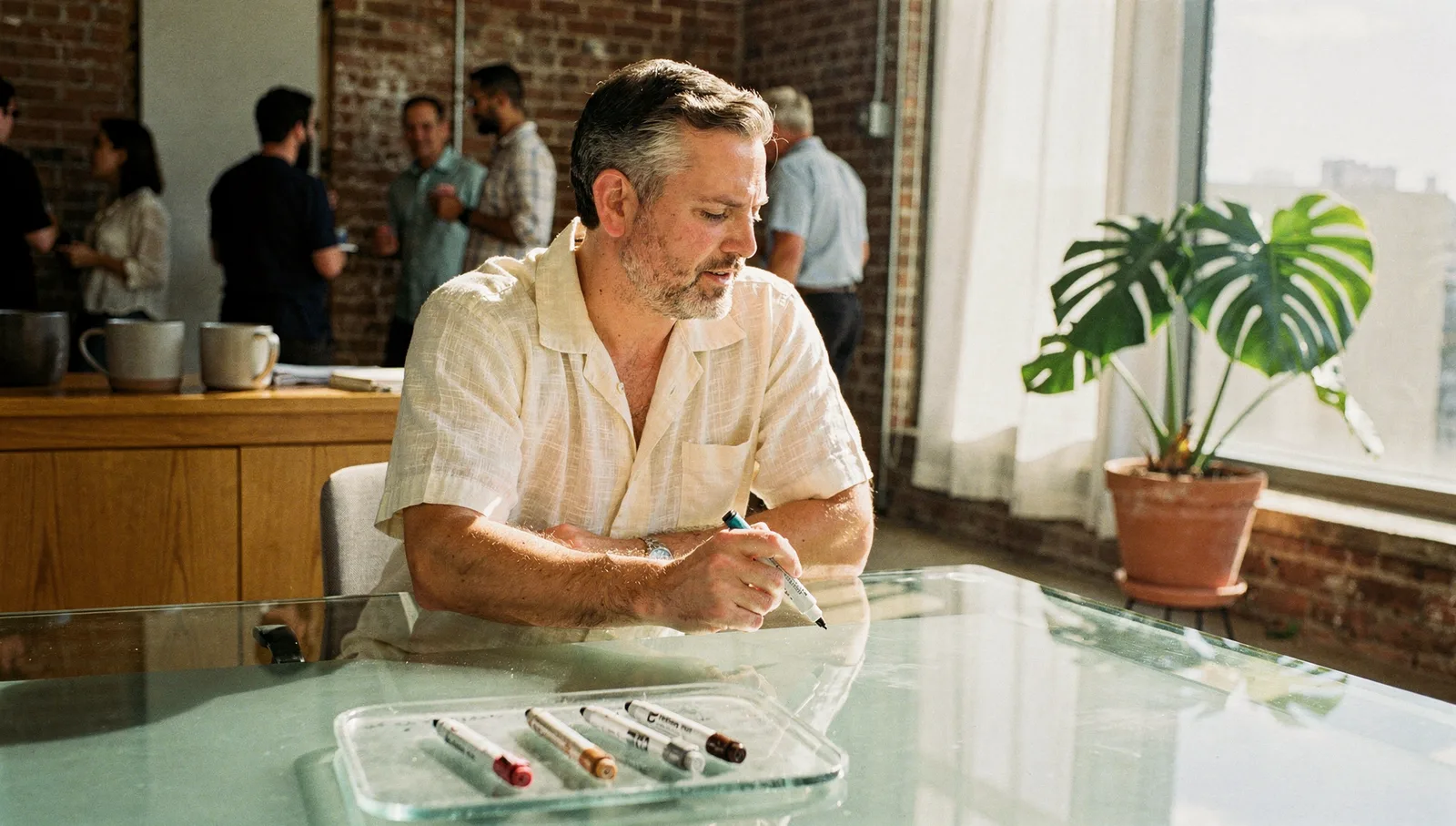 Man in a guayabera leaning over a glass meeting table uncapping a marker in a bright agency office.