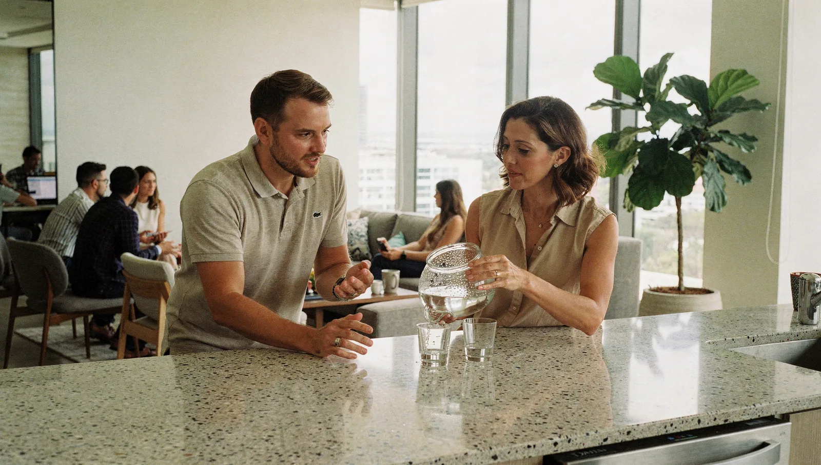 Two colleagues talking at a sunlit office kitchen counter while a woman pours water from a carafe into a glass.