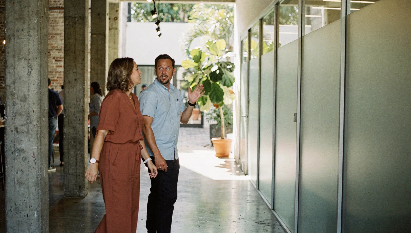 Two colleagues talk while walking down a sunlit, modern warehouse hallway with concrete pillars and a potted tree.