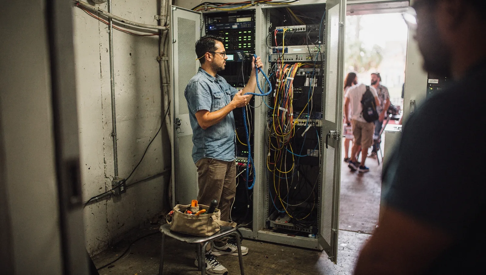 A technician inspects a blue network cable while troubleshooting inside a cramped server closet.