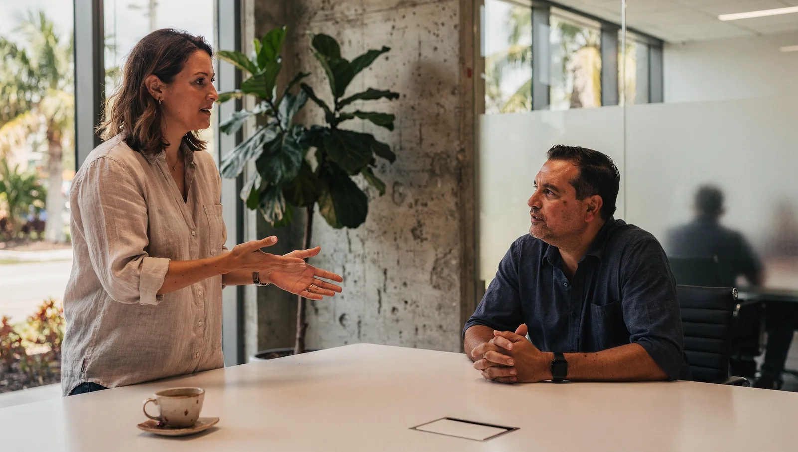 A marketing strategist speaks with a business owner in a sunlit conference room with a fiddle-leaf fig tree.